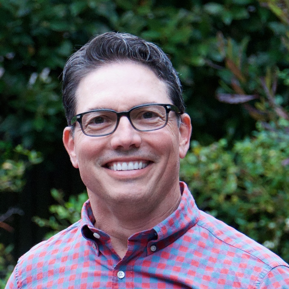 A man with rosy complexion, dark grey hair and glasses smiles at the camera. He is wearing a red and blue checkered shirt and stands against a backdrop of greenery.